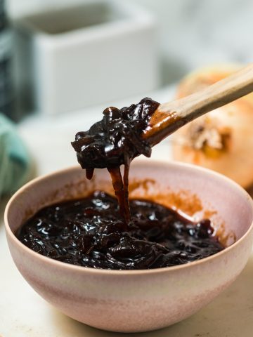 A pink porcelain bowl filled with dark brown balsamic onion jam. A wooden spoon full of the jam hovers over the bowl.