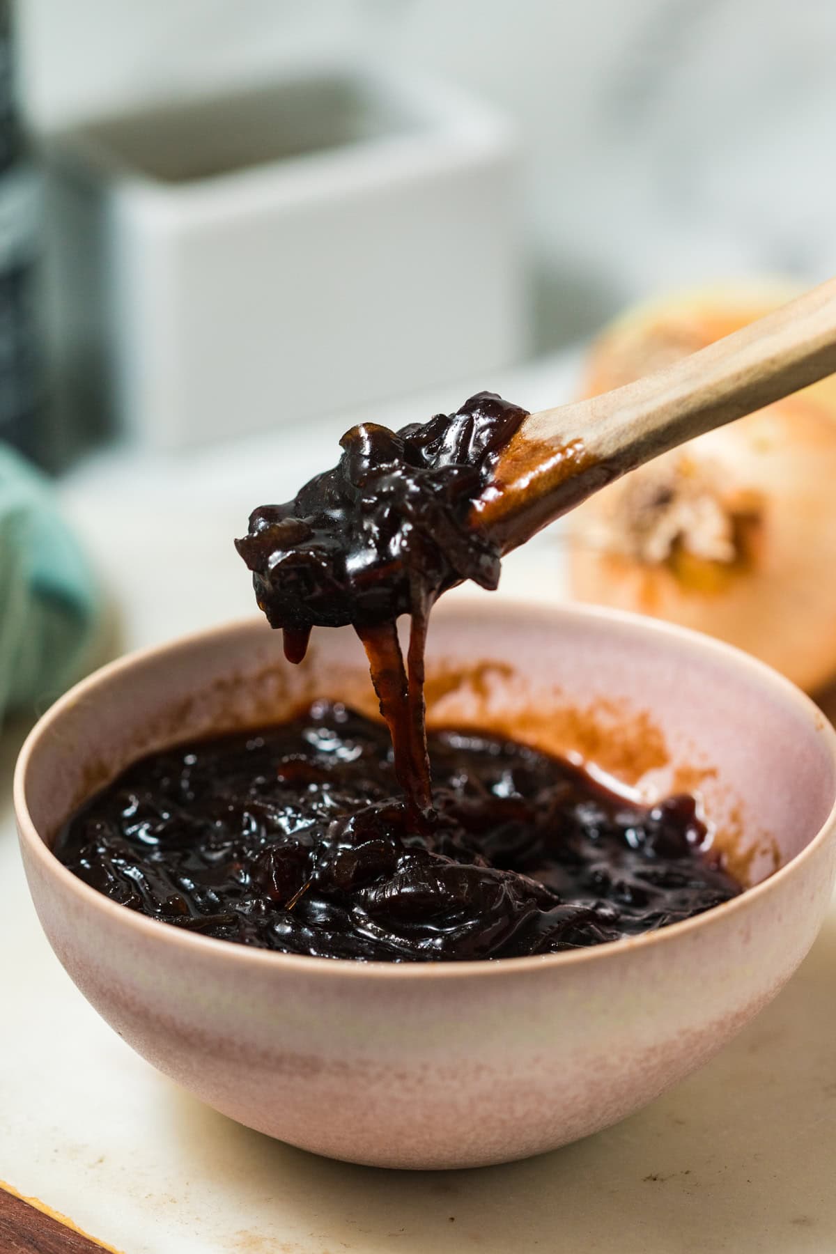 A pink porcelain bowl filled with dark brown balsamic onion jam. A wooden spoon full of the jam hovers over the bowl. 