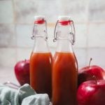 Two glass bottles of boiled apple cider in front of a tile wall. The bottles are surrounded by red apples.