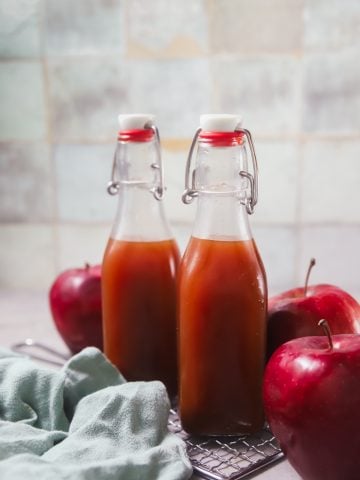 Two glass bottles of boiled apple cider in front of a tile wall. The bottles are surrounded by red apples.