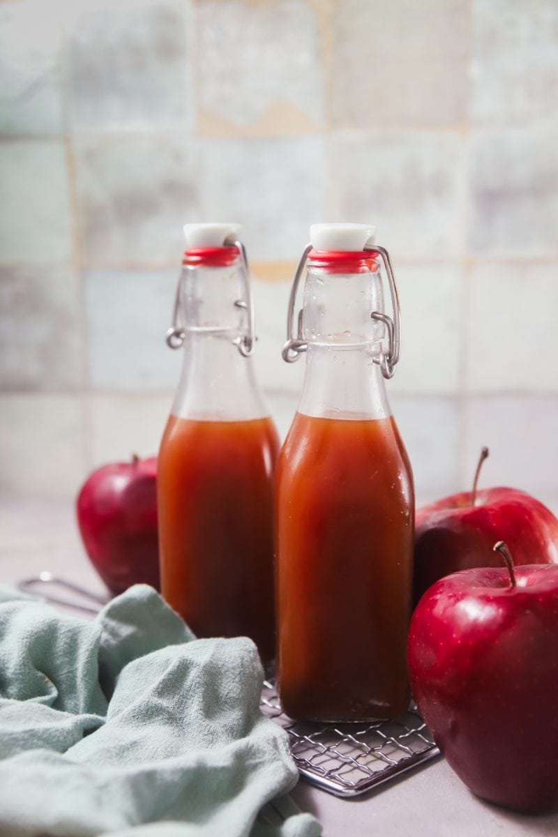 Two glass bottles of boiled apple cider in front of a tile wall. The bottles are surrounded by red apples.