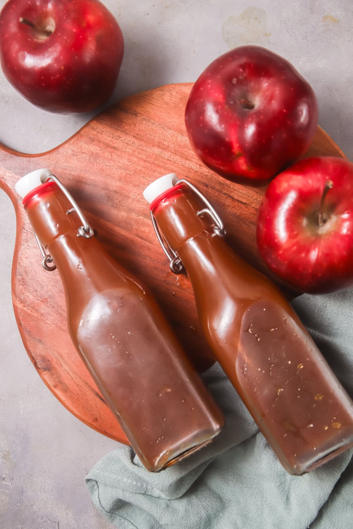 Overhead view of two glass bottles of boiled cider laying on a wooden serving board surrounded by apples. 