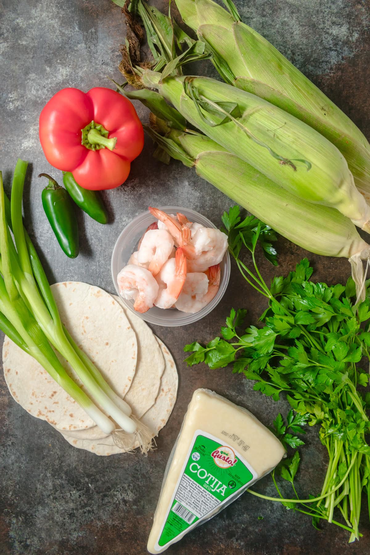 Flatlay image of ingredients needed to make shrimp and corn tacos, including red bell pepper, cilantro, scallions, jalapenos, and cotija cheese, arranged on a dark grey surface. 