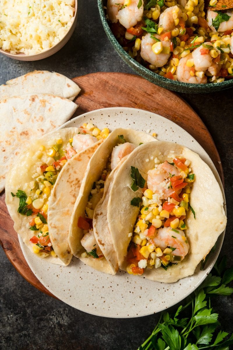 An overhead view of three shrimp and corn tacos. There's a bowl of Mexican steet corn inspired corn salsa and a dish of cotija cheese peeking into the frame. 